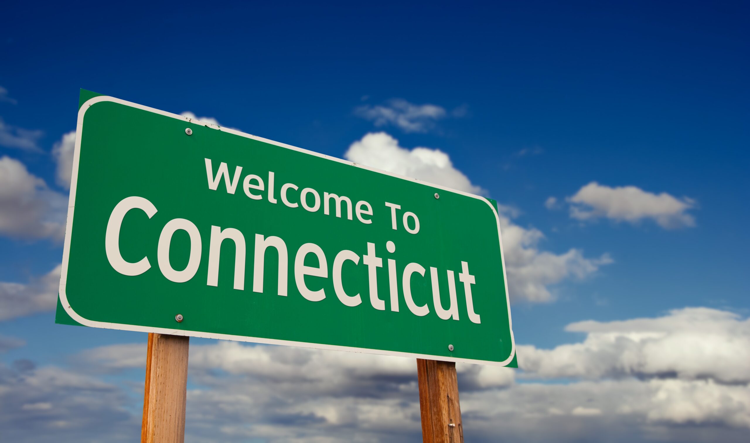 Green road sign reading 'Welcome to Connecticut' against a blue sky with scattered clouds (tilted on wooden posts).
